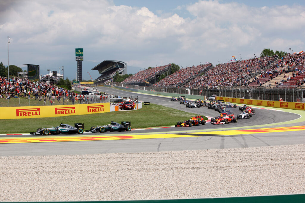 Formel-1-Autos beim Start am Circuit de Barcelona-Catalunya, mit vollen Zuschauertribünen im Hintergrund.