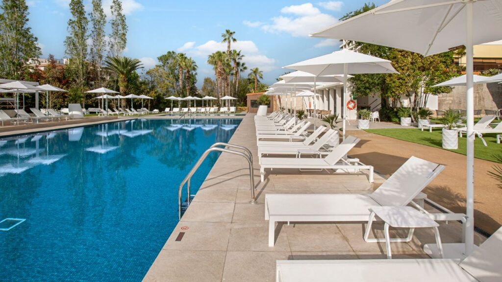 Breiter Pool mit weißen Sonnenliegen und Sonnenschirmen, umgeben von Palmen und Garten.