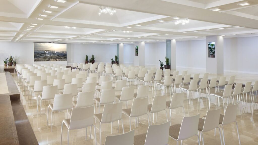 Großer heller Konferenzraum mit Reihenstühlen, heller Marmorfläche und Leinwand.