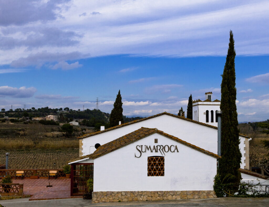 Weißes Gebäude des Weinguts Sumarroca mit Zypressen und umliegender Weinlandschaft unter leicht bewölktem Himmel.